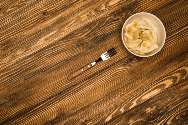Вид зверху смачні вареники зі смаженою цибулею в мисці на дерев'яному столі з виделкою — стокове фото