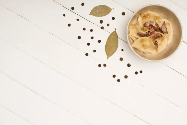 Top view of delicious varenyky with cracklings in bowl near bay leaves and black peppercorns on white wooden table — Stock Photo