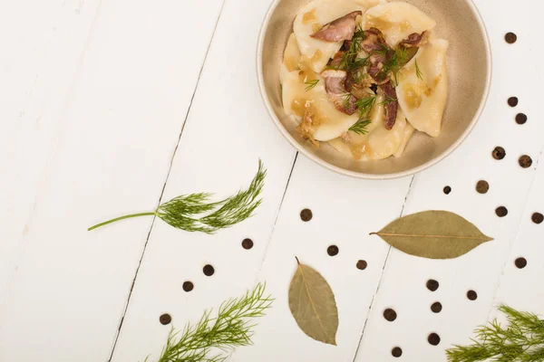 Draufsicht auf köstliche Varenyky mit Knistern und Dill in Schüssel in der Nähe von Lorbeerblättern und schwarzen Pfefferkörnern auf weißem Holztisch — Stockfoto