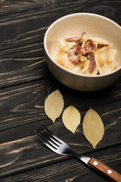 Délicieux varenyky avec craquelins dans un bol près de feuilles de laurier et fourchette sur table en bois noir — Photo de stock