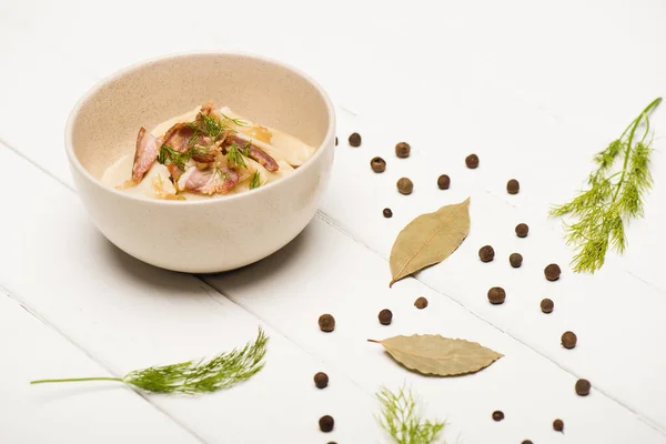 Delicious varenyky with cracklings and dill in bowl near bay leaves and black peppercorns on white wooden table — Stock Photo