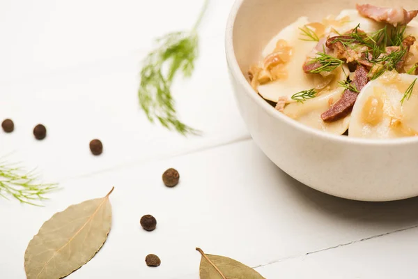 Foyer sélectif de délicieux varenyky avec des craquelins et de l'aneth dans un bol près des feuilles de laurier et des grains de poivre noir sur une table en bois blanc — Photo de stock