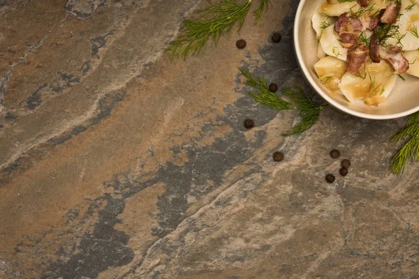 Vue de dessus de délicieux varenyky avec des craquelins dans un bol près de l'aneth et des grains de poivre noir sur la surface de la pierre — Photo de stock