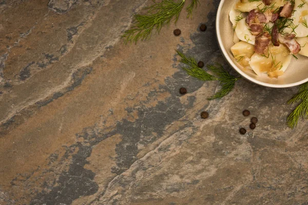 Top view of delicious varenyky with cracklings in bowl near dill and black peppercorns on stone surface — Stock Photo