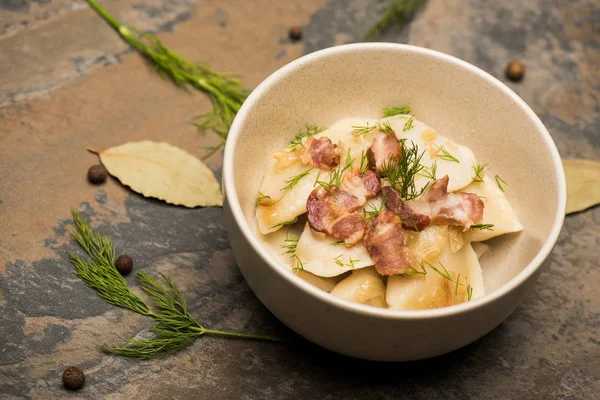 Delicious varenyky with cracklings and dill in bowl near bay leaves and black peppercorns on stone surface — Stock Photo