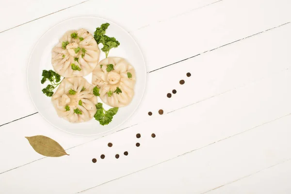 Top view of delicious Khinkali with cilantro on plate near bay leaf and black peppercorns on white wooden table — Stock Photo