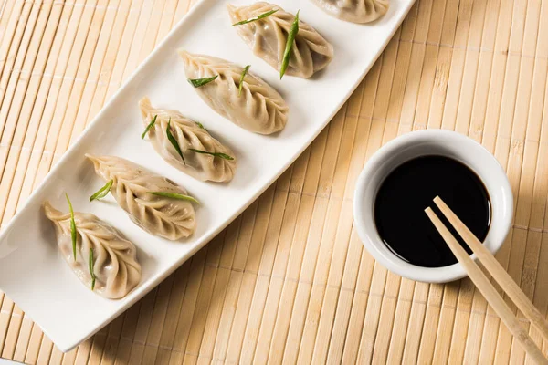 Вид на вкусные китайские вареные пельмени на тарелке рядом с палочками и соевым соусом на бамбуковом коврике — стоковое фото