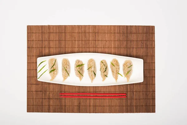 Вид сверху на вкусные китайские вареные пельмени на тарелке рядом с палочками для еды на бамбуковом коврике на белом фоне — стоковое фото