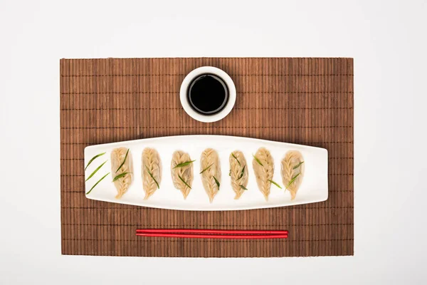 Вид на вкусные китайские вареные пельмени на тарелке рядом с палочками и соевым соусом на бамбуковом коврике на белом фоне — стоковое фото
