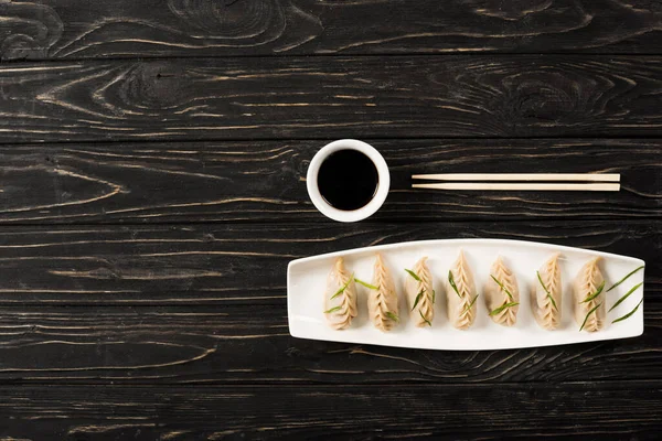 Vue du dessus de délicieuses boulettes bouillies chinoises sur assiette près des baguettes et de la sauce soja sur table en bois noir — Photo de stock