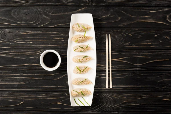 Vue du dessus de délicieuses boulettes bouillies chinoises sur assiette près des baguettes et de la sauce soja sur table en bois noir — Photo de stock