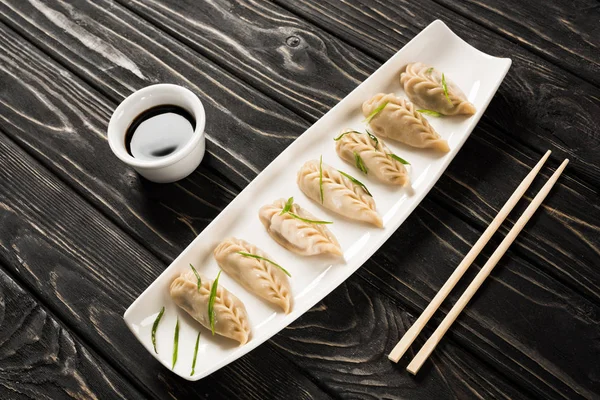 Вкусные китайские вареные пельмени на тарелке рядом с палочками и соевым соусом на черном деревянном столе — стоковое фото