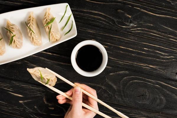 Cropped view of woman eating delicious Chinese boiled dumpling with chopsticks and soy sauce at black wooden table — Stock Photo