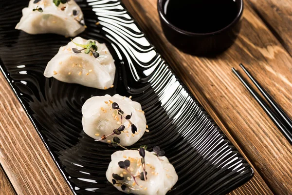 Крупный план вкусных китайских варёных пельменей на тарелке возле палочек для еды и соевого соуса на деревянном столе — стоковое фото