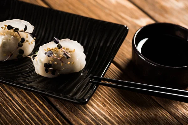 Крупный план вкусных китайских варёных пельменей на тарелке возле палочек для еды и соевого соуса на деревянном столе — стоковое фото