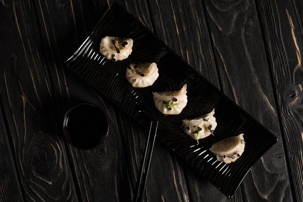 Вид на вкусные китайские вареные пельмени на тарелке рядом с палочками и соевым соусом на черном деревянном столе — стоковое фото