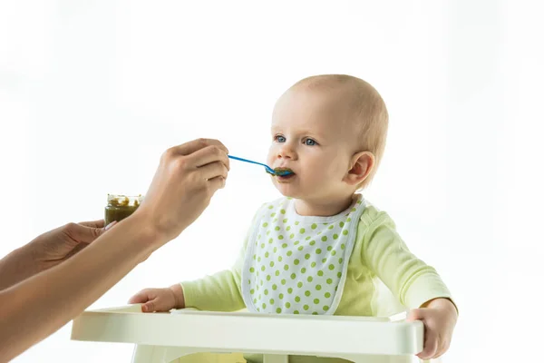 Madre con barattolo di nutrizione vegetale per bambini e cucchiaio che si nutre di bambino sulla sedia di alimentazione isolata su bianco — Foto stock