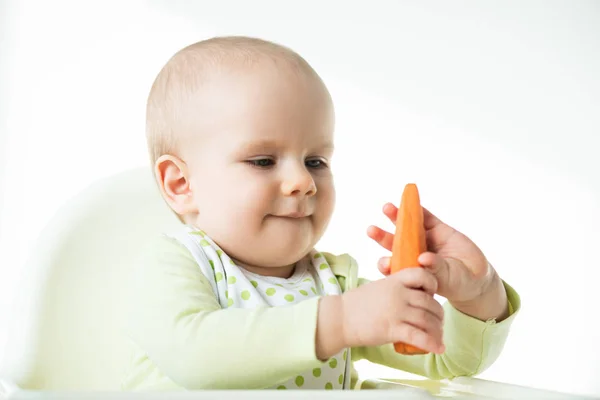 Liebenswertes Baby, das Karotte hält, während es auf einem Fütterungsstuhl sitzt, isoliert auf weißem Grund — Stockfoto