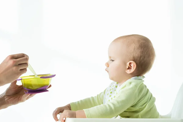 Vista lateral de la madre sosteniendo tazón de la nutrición del bebé y cuchara cerca del bebé en la silla de alimentación sobre fondo blanco - foto de stock