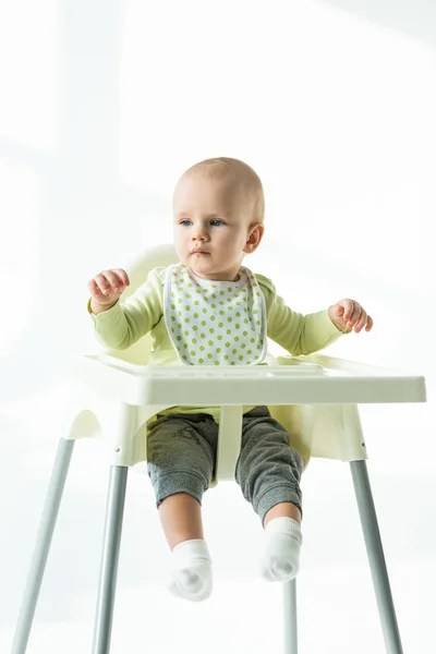 Bebé sentado en la silla de alimentación y mirando hacia otro lado sobre fondo blanco - foto de stock