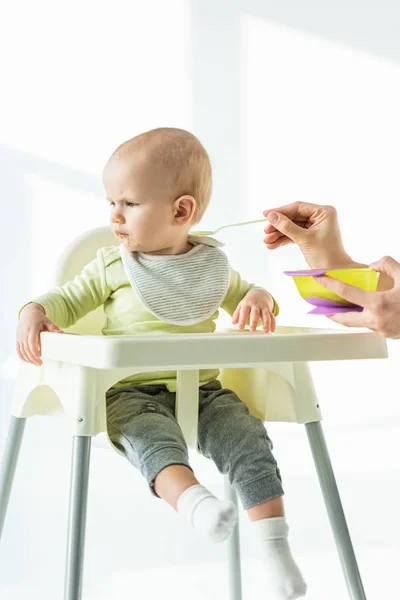 Bebê na cadeira de alimentação olhando para longe perto da mãe com tigela de nutrição do bebê e colher no fundo branco — Fotografia de Stock