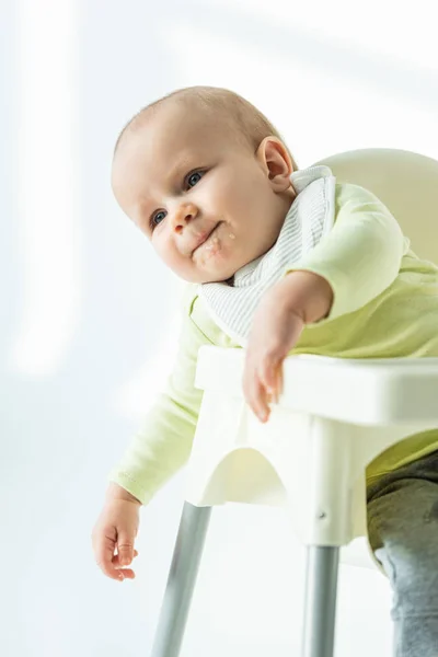 Lindo bebé con la boca sucia en puré sentado en la silla de alimentación sobre fondo blanco - foto de stock