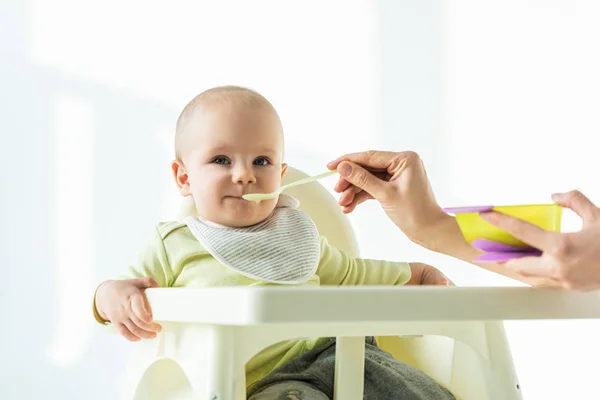 Foco seletivo da mãe com purê de alimentação do bebê na cadeira de alimentação no fundo branco — Fotografia de Stock