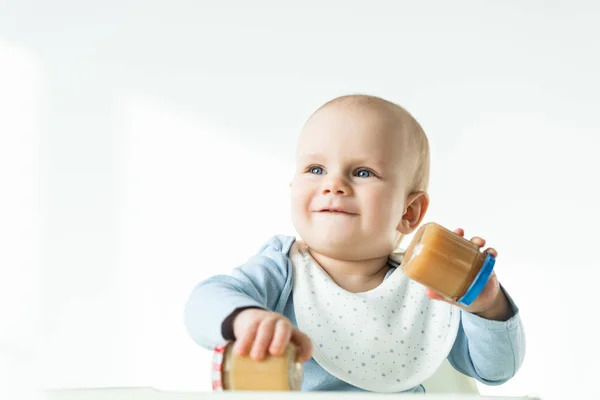 Foco seletivo de bebê segurando frascos de frutas nutrição do bebê e sorrindo enquanto sentado na cadeira de alimentação no fundo branco — Fotografia de Stock