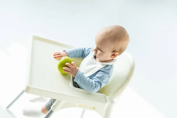 Vista de ángulo alto del bebé niño sosteniendo manzana verde mientras está sentado en la silla de alimentación sobre fondo blanco - foto de stock