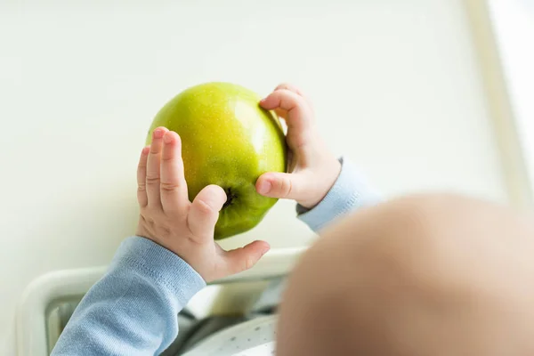Overhead view of baby holding green apple while sitting on feeding chair — Stock Photo