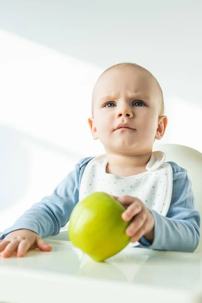 Selektiver Fokus des nachdenklichen Jungen mit grünem Apfel, während er auf einem Fütterungsstuhl auf weißem Hintergrund sitzt — Stockfoto