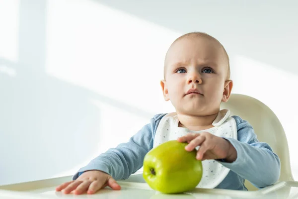 Mignon bébé garçon touchant pomme verte sur la table de la chaise d'alimentation sur fond blanc — Photo de stock