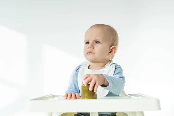 Lindo bebé niño sosteniendo tarro de puré de verduras y mirando hacia otro lado en la silla de alimentación sobre fondo blanco - foto de stock