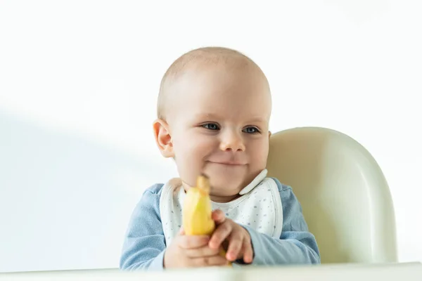 Selective focus of smiling baby holding banana while sitting on feeding chair on white background — Stock Photo