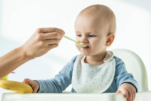 Mother feeding baby son while holding banana on table of feeding chair on white background — Stock Photo
