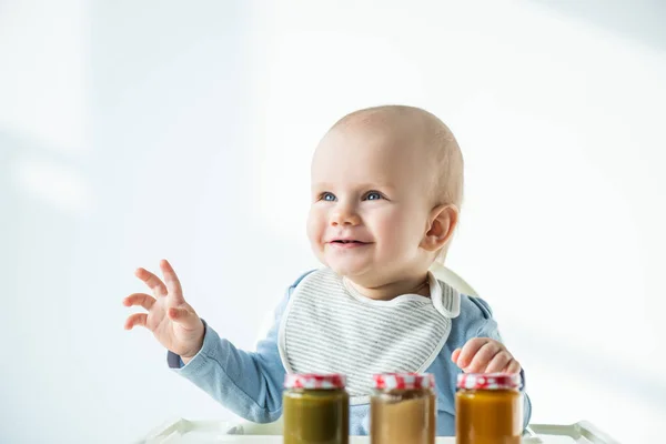 Foyer sélectif de bébé garçon souriant loin près des pots de nutrition bébé sur la table de chaise d'alimentation sur fond blanc — Photo de stock