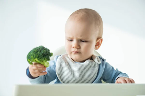 Selective focus of baby holding piece of broccoli while sitting on feeding chair on white background — Stock Photo