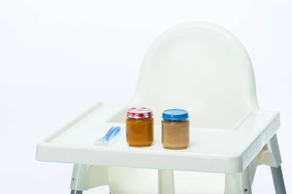 Jars of baby food and spoon on table of feeding chair isolated on white — Stock Photo
