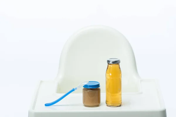 Jar of baby food with spoon and bottle of apple juice on table of feeding chair isolated on white — Stock Photo