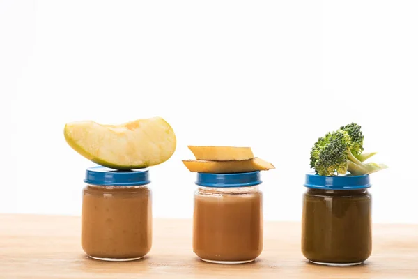 Frascos de alimentos para bebés con trozos de manzana, brócoli y rodajas de plátano en la superficie de madera aislada en blanco - foto de stock