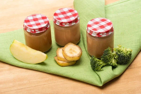 Frascos de nutrição infantil preparada com pedaços de maçã, brócolis e fatias de banana no guardanapo no fundo de madeira — Fotografia de Stock