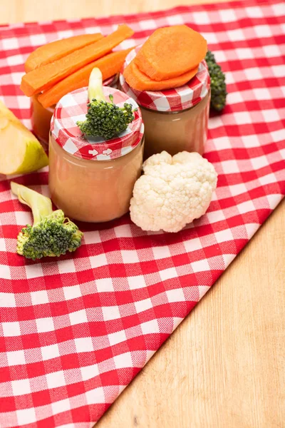 Jars of baby nutrition with fresh apple, carrot and broccoli on checkered napkin on wooden surface — Stock Photo