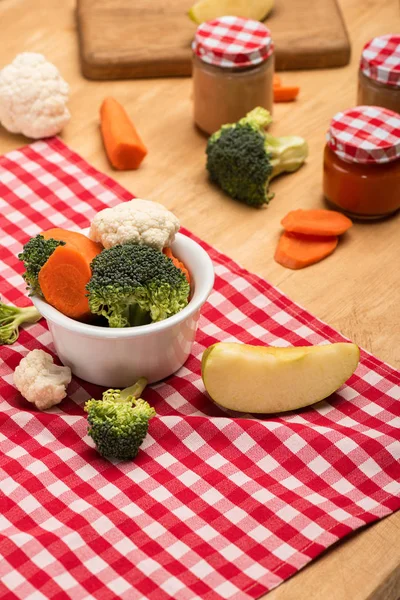 Organic vegetables and apple on napkin near jars of baby food on wooden background — Stock Photo