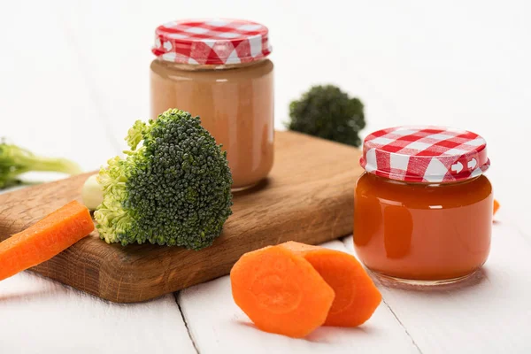 Close up view of fresh broccoli, carrot and jars of baby food on cutting board on white wooden background — Stock Photo