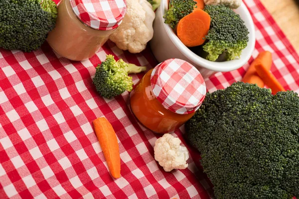 Vista de alto ângulo de brócolis, cenouras e couve-flor perto de frascos de comida para bebês na toalha de mesa em fundo de madeira — Fotografia de Stock