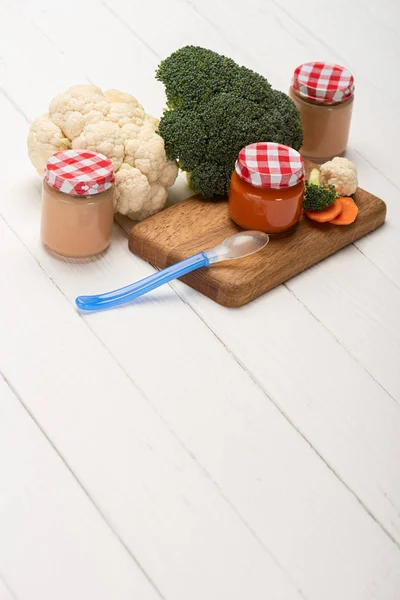 Nutrição do bebê em frascos com legumes maduros e colher na tábua de corte na superfície de madeira branca — Fotografia de Stock