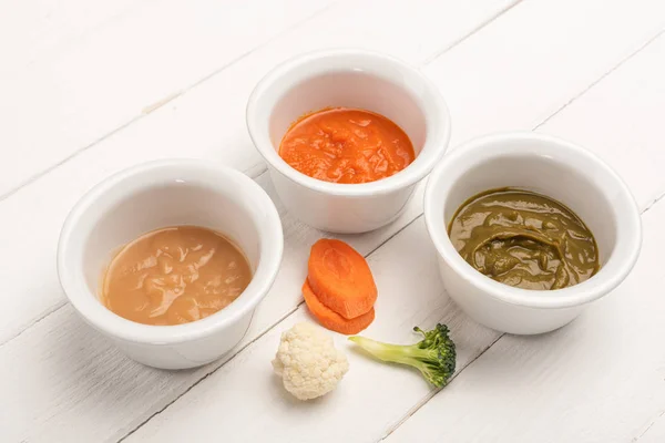 Bowls with baby nutrition with carrot slices and pieces of cauliflower and broccoli on white wooden background — Stock Photo