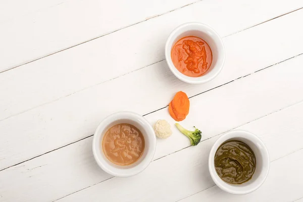 Top view of carrot slices, pieces of cauliflower and broccoli near bowls with baby food on white wooden surface — Stock Photo