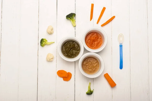 Top view of baby nutrition in bowls with pieces of vegetables and spoon on white wooden surface — Stock Photo