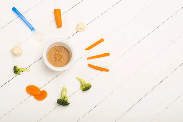 Вид зверху миски з кольоровим дитячим харчуванням біля шматочків овочів і ложки на білому дерев'яному фоні — стокове фото